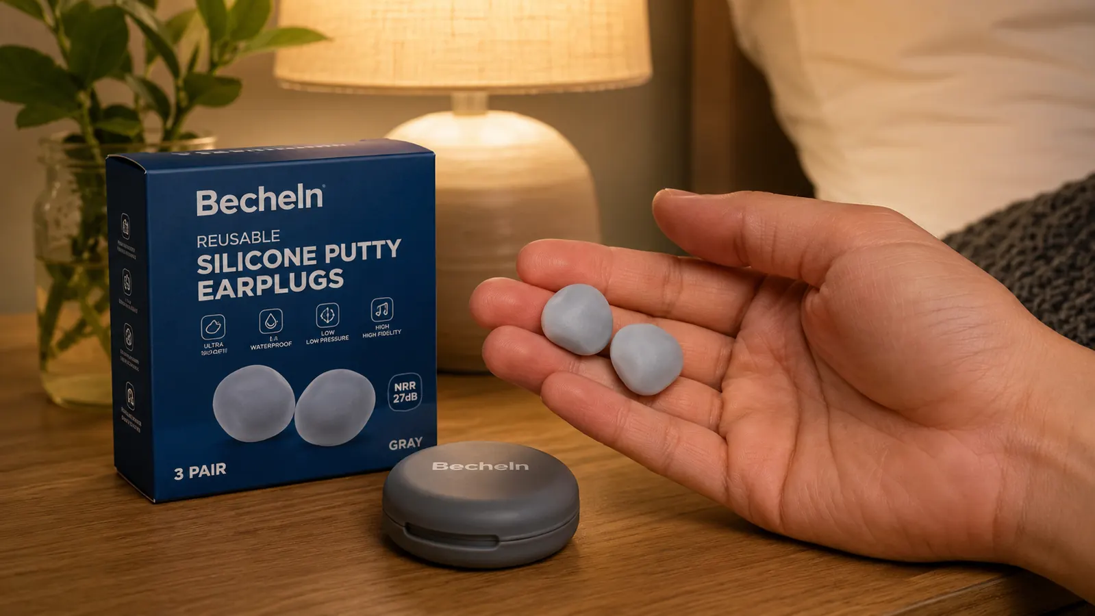 Hand holding a pair of moldable silicone earplugs above a nightstand with a lamp and glass of water